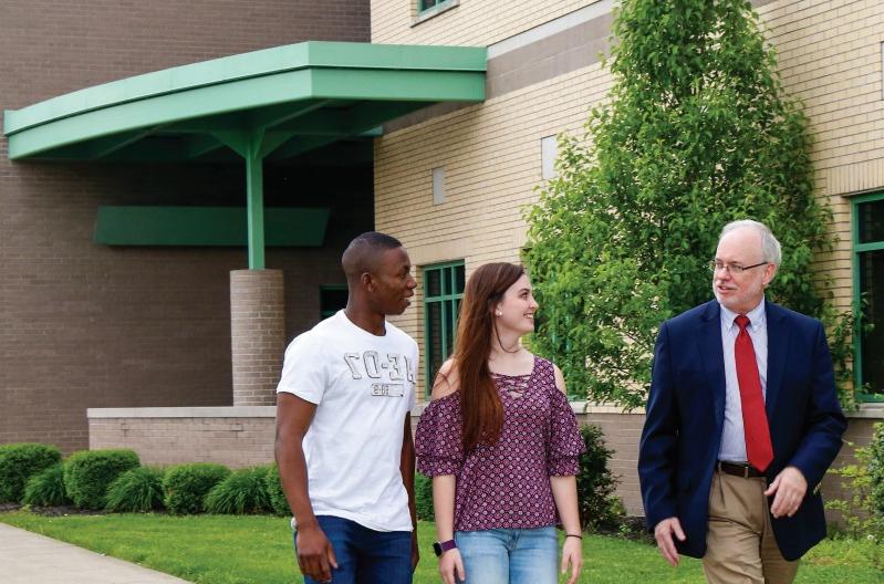 三个人 are walking and conversing outside a building with green trim. A man in a suit and tie is accompanied by two young adults, 一个穿着有图案的衬衫和牛仔裤, 另一个穿白t恤.